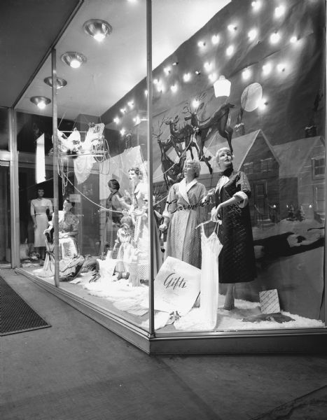 Carmen's Christmas window at 9 South Pinckney Street, featuring female mannequins wearing the latest fashions for winter, including dresses, robes ("house coats") and pajamas. The mannequin closest to the front holds a spaghetti strap nightgown on a hanger. The window display shows a snowy scene with a simple metal sleigh, loaded with wrapped packages and clothing, that is attached by rope to reindeer flying past village homes and a full moon. Fake snow covers the flooring around the feet and edges of the display. A sign at the front reads: "Gifts."