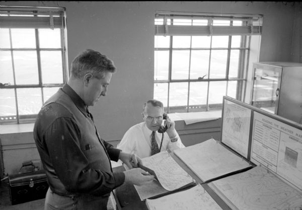 Chief of weather operations, L.A. Joos, is on the telephone, while W.C. Williamson is standing at a slanted desk checking one of several weather maps. They are working in the Truax Field control tower. The view outside the two windows looks out towards the horizon.