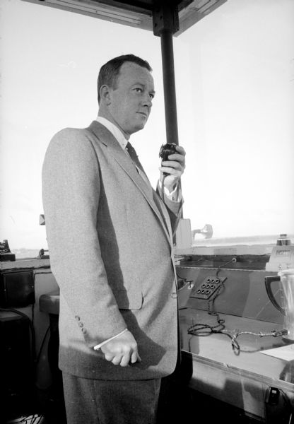 Edward C. Waffle, in charge of the tower for the Air Force, is talking with a pilot from the Truax Field control tower by speaking into a handheld radio transmitter while looking out of the observation window.