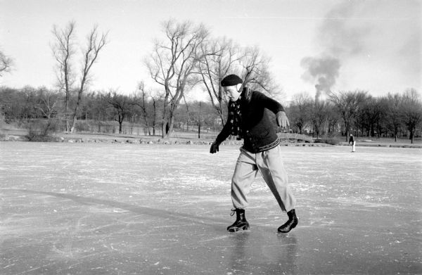 Dave Halls is trying out new ice skates he received for Christmas at Tenney Park pond, which is used as a Madison outdoor rink in winter. With ankles wobbly on the blades, he steadies himself with arms out. He is dressed in gloves, ear muffs, scarf and sweater. Another skater and the Tenney Park bridge are in the background. A plume of smoke is rising from east side industry beyond the park's trees.