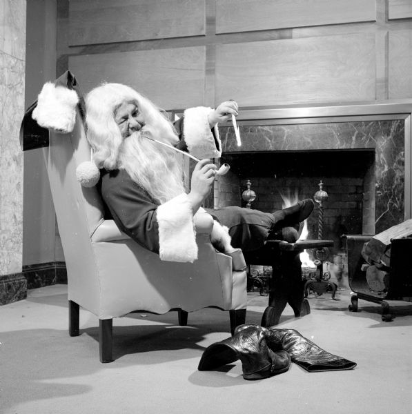 Man dressed as Santa Claus sitting in front of a fireplace in an armchair, lighting his pipe with a candle. His pom-pom hat is hanging over the back of the chair, and his boots are lying on the floor beside him, while he is resting his feet on a table to warm them by the fire.