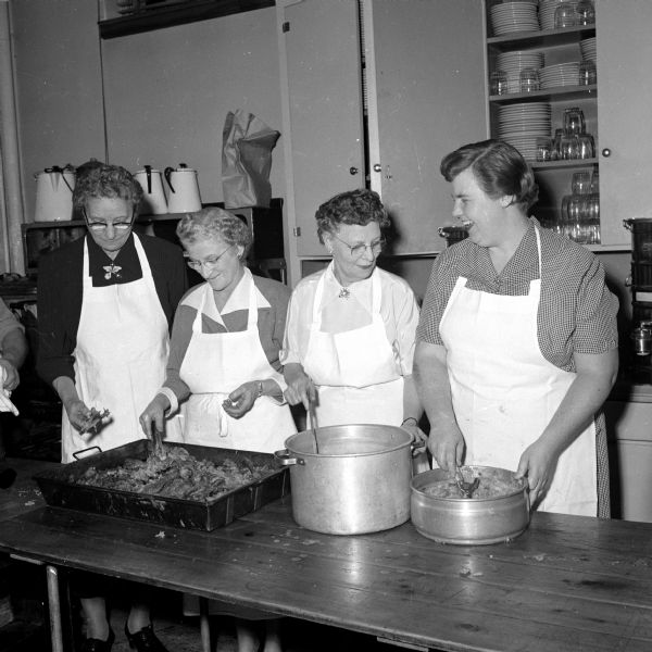 Cooks preparing food for the Rennebohm Drug Store annual Christmas party in the kitchen of the Catholic Community Center. Left to right are: Ella Claus, Katherine Marty, Mabel Rider, and Helen Fossen. They all wear white aprons over nice dresses, attending to various sized pots.