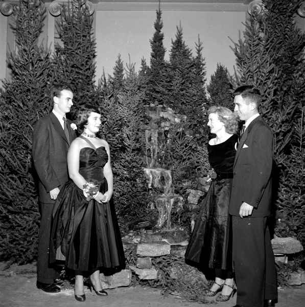 The annual Christmas Charity Ball held at the Loraine Hotel. Four members of the decorations committee survey their handiwork. Left to right are: Reed Coleman, Muff Hobbins, Juliette Gerke, and John Haydon. They are all in formal attire, men in suits and ladies in gowns and high heel shoes, standing before a thick forest of cut pine trees and a waterfall installation inside the Crystal Ballroom. 