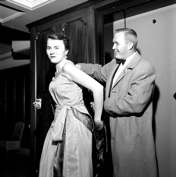 The annual Christmas Charity Ball held at the Loraine Hotel. Joan Fagan, general chairperson for the ball, is helped with her coat by Denny Cosgrove. She is wearing a strapless gown with a bow at the waist and he is in long wool overcoat. 