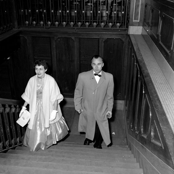 The annual Christmas Charity Ball held at the Loraine Hotel. Walking up the stairs to the Crystal Ballroom are Mary Ann Hand, who was in charge of freshman invitations, and her escort, Jim Fagan. They are both in formal attire, he in black-tie and she a glittering gown with white gloves and choker necklace. 