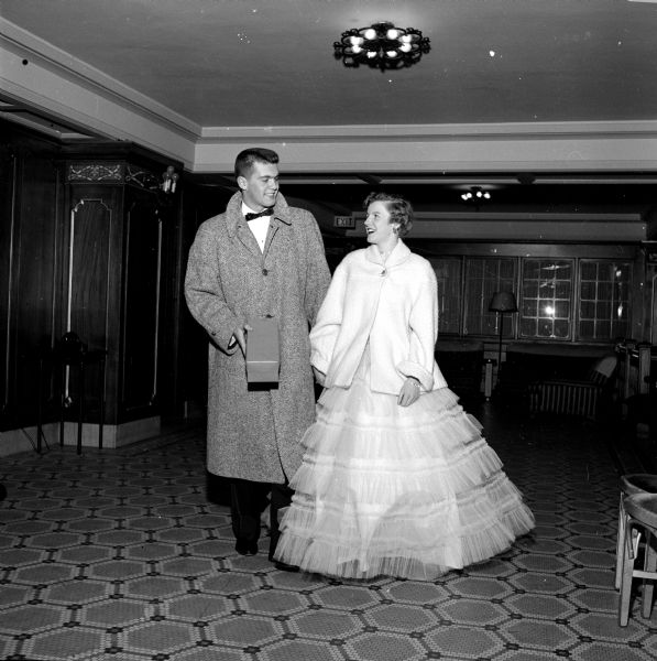 The annual Christmas Charity Ball held at the Loraine Hotel. Tom Stephan holds a box containing a corsage for his dance date, Jean McFarland. Standing on a tile-mosaic floor, they are posing in formal attire: he is wearing black-tie and she is wearing a cascade tiered gown. 