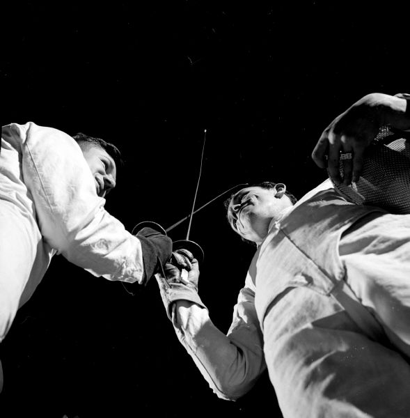 View from below of Jack Heiden and Bruce Backmann of the University of Wisconsin fencing team crossing foils in the time-honored salute at the beginning of a fencing match against Shorewood. The man on the right is holding his mask under his left arm.