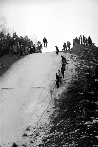 View up hill towards Don West, of the host Blackhawk Ski Club, who has just left the jumping scaffold, competing in the Class C category. Spectators are along the upper and lower part of the jump. 