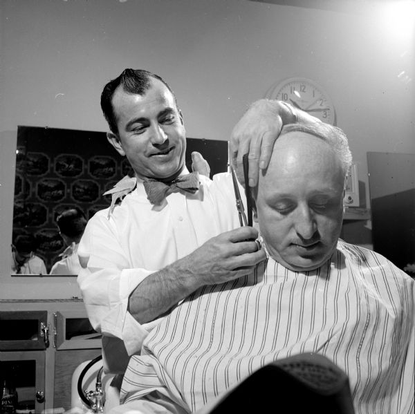 Barber Roy Brumley giving Dan Seligman a haircut while his two pet parakeets are perched on each of his shoulders, at the Belmont Hotel barbershop. Seligman is draped in a striped apron and Brumley is wearing a bow-tie and white shirt. 
