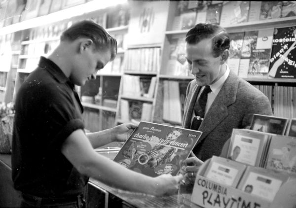 Record shop employee Charles Lunde (right) explaining the merits of Charlie Ventura's septet to customer Richard Mallum, who is admiring the cover of the vinyl record. Other 33rpm records line the back wall, and 45s are sitting in a small rack on the counter top.