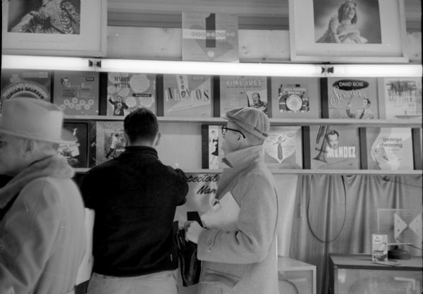 Customer John Salzer (left) looking over an album with George Dasson. Other 33rpm vinyl records are displayed on the wall, and framed photographs of musical artists are sitting on top of the rack. 