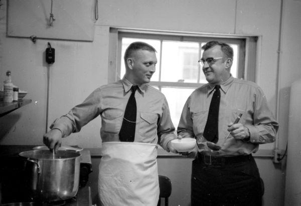 Harold Hansen (left) and Fred Manthe making chili for the firemen's dinner. Hansen is standing next to the stove stirring a deep pot with one hand, and is holding a white bowl in the other hand. Manthe is holding a ladle. 