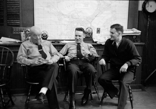 Chatting about previous fire fighting experiences are (from left to right): Captain Arthur Wilcox, Firemen Russell Mani, and Wilbur Wright. They are sitting in an office at the fire station in front of a large map of Madison tacked to the wall behind them.