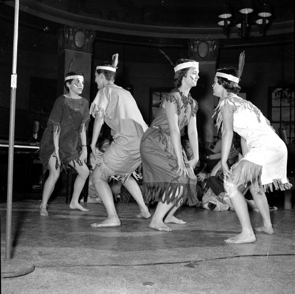 Four sorority sisters performing an Indian "war dance" on behalf of their candidate for prom queen, Doris Sickert from Wauwatosa, at the rally at the Memorial Union Rathskellar. The performers are, left to right: Mary Jo Perreault, Marilyn Fullerton, Kathy Kading, and Aliee Jones. They are wearing stereotypical dresses made to look like fringed buckskin, are barefooted, and have a single tall feather at the back of their heads. 