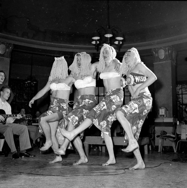 Four male "chorus girls" dancing on behalf of their prom queen candidate, Gloria Robbins of Oshkosh, at the rally at the Memorial Union Rathskellar. The performers are, left to right: Bill Haanen, Dick Awe, Don Dedow van Dyne, and Dick Traegger. Barefooted, they are wearing strapless bras, tropical floral knee-length skirts, and white-mops as wigs. 