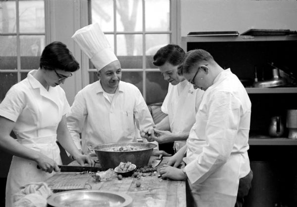 Instructor E.L. Heiser looking on as students Jean Burik, Bob Knoelke, and Delwin Henriksen are preparing a salad in the cooking school at Madison Vocational School. The group is wearing white aprons and chef shirts.