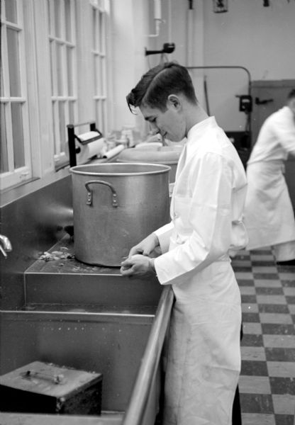 Student Harold Loskot peeling potatoes in the cooking school at the Madison Vocational School. Dressed in a white apron and chef's shirt, he is standing at a stainless steel sink by a tall pot. 