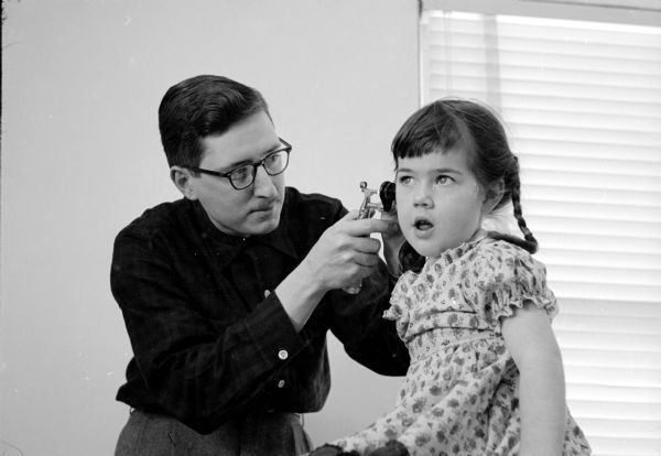 Staff member Dr. M.O. Kepler looking into the ear of little Sara Willey at the Cuba City Hospital and Clinic. Her hair is braided. 