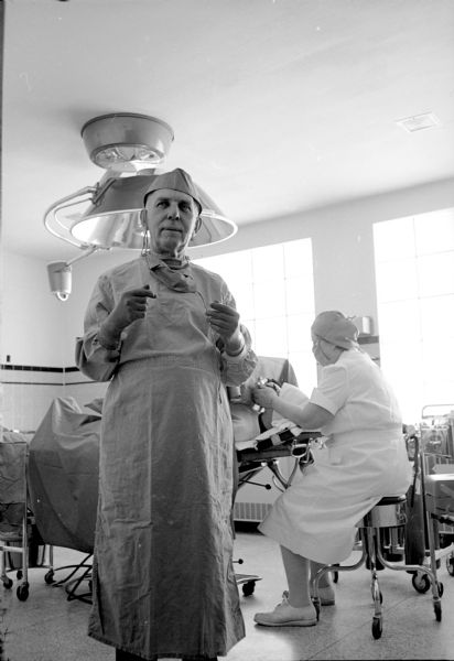 Surgeon Dr. K.K. Rossmann preparing for surgery at the Cuba City Hospital and Clinic. He is scrubbed up for the operating room in his gloves, hat and robe. A nurse is sitting behind him administering anesthesia to a patient on the table. 