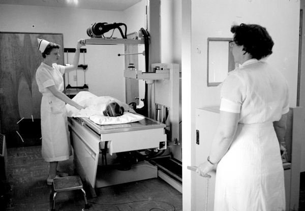 Nurse Marie Holt adjusting the new X-ray machine over a patient lying on the table, while Selma Haase, in the right foreground, is waiting to take a picture.