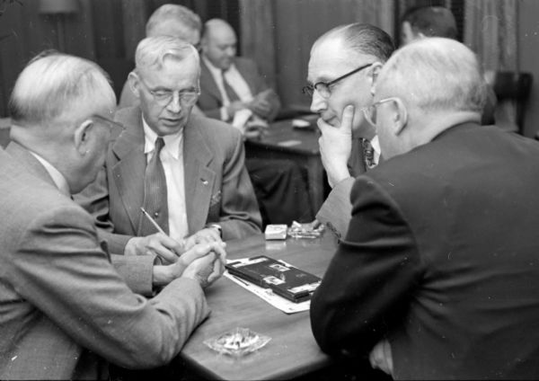 Adding up the scores at the men-only bridge tournament at the City YMCA are Professor M. Starr Nichols, Dr. R.T. Vogel, Herbert Hawkinson, and H.F. Loettler. Three men are looking at the score tallier, their elbows on a table by glass ashtrays and a pack of cigarettes.