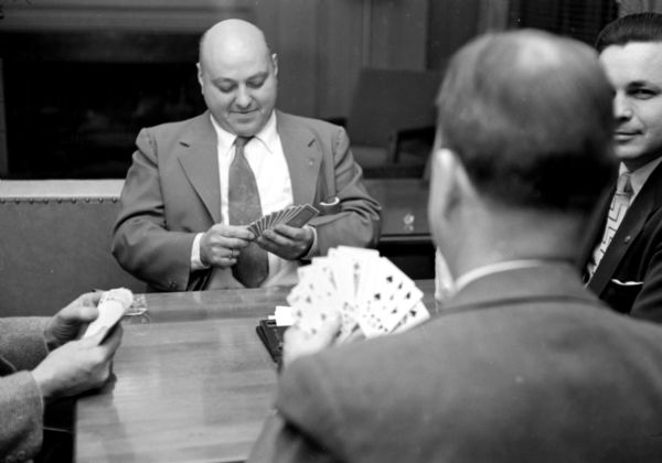 A.R. Sergenian and Tester Bakken are partners at the men-only bridge tournament at the City YMCA. One of the four players' cards is in the foreground. On the left a man's hands holding cards is obscuring a glass ashtray on the corner of the table. 