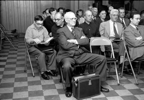 Club treasurer, Charles P. Ingold, sitting among a crowd at the Madison Stamp Club meeting at the City YMCA. Behind him, Edgar Phillips is showing a philatelic album to Arthur Duve.