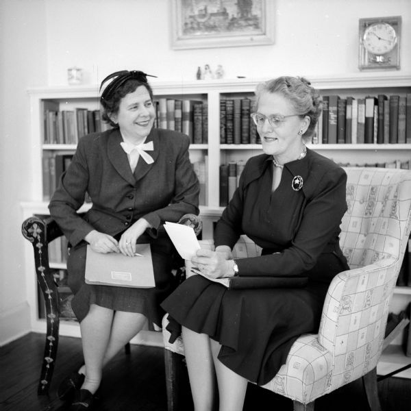 Isabel La Follette (right) and probably Deborah Sherman, are discussing the Woman's Service Exchange, which Mrs. La Follette started at the Madison Vocational and Adult School. It was designed to unite older employable women, who wanted to work, with employers, who needed women workers. Mrs. Sherman was the Executive Secretary of the Exchange Committee. They are both sitting in armchairs in front of a book shelf, clock and framed painting.