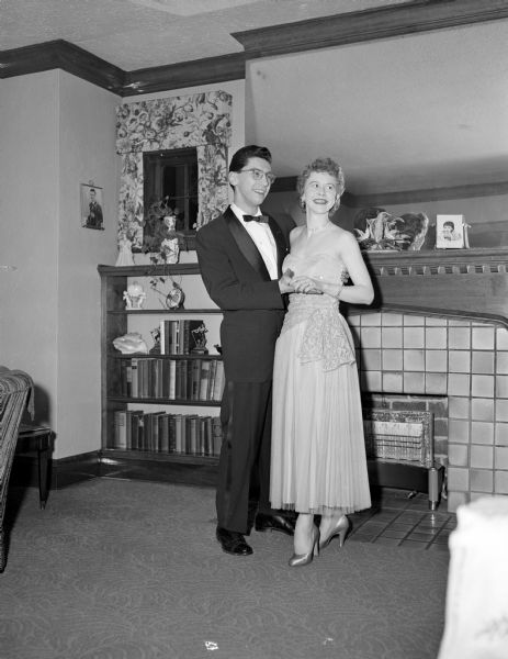 Photographer Ed Stein and his wife, Donna, in formal dress before attending the University of Wisconsin prom at the Memorial Union. He is in a tuxedo and she in a strapless ballgown. They are standing holding hands before a fireplace in a private home. Above the fireplace mantel is a mirror, and beside it is a bookshelf.
