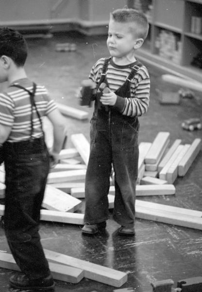 Randy Kross banging blocks together at play time. He is wearing coveralls, and many boards are strewn on the floor at his feet.
