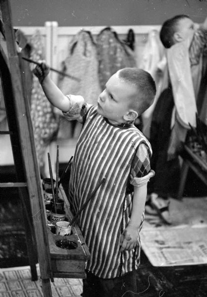 Levi Wood painting at an easel. His sleeves are rolled up and he is wearing a smock. In the background is a wall of coats hanging from hooks, and another boy painting at an easel.