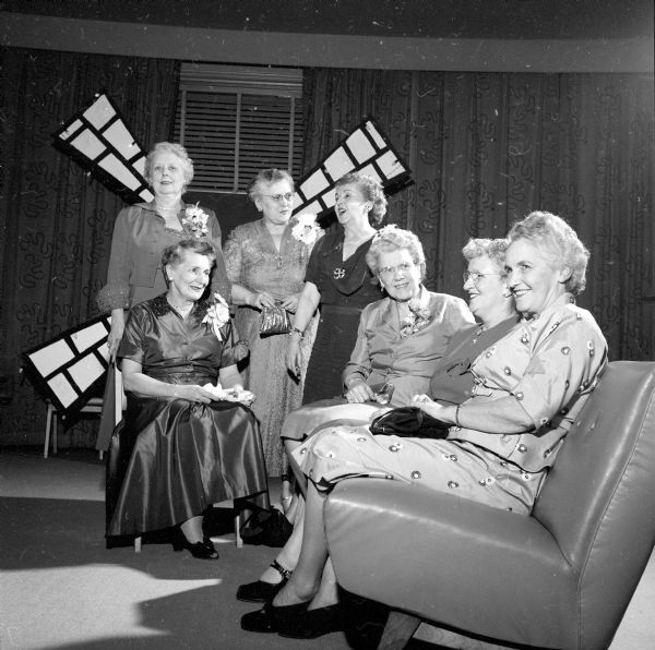 Chatting at the Panhellenic Ball in the Great Hall of the Memorial Union are sorority housemothers. Seated are Mrs. Ruth Gauchat, Mrs. Nyla Lowell, Mrs. Kenneth Petton, and Mrs. Sarahbelle Slemmons; and standing in front of a windmill, Mrs. William C. Smith, Mrs. E.J. Link and Mrs. O.R. Riday. They are all formally dressed, and some of them are wearing corsages.