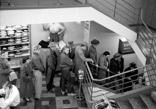 View looking down at a line of men and women waiting to buy tickets to Milwaukee Braves baseball games at the branch ticket office located in the basement of The Hub clothing store in downtown Madison.