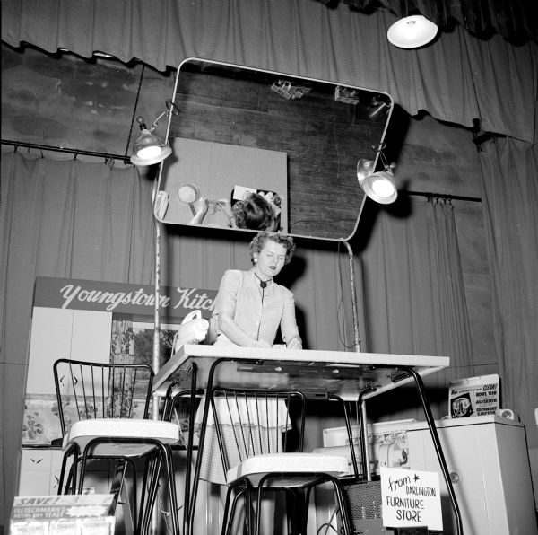 Youngstown Kitchens appliance show sponsored by the Darlington Furniture Store held in the Darlington High School auditorium. A woman is demonstrating new kitchen appliances on a table, and a mirror is set up above and behind her so the audience can see what she is doing.
