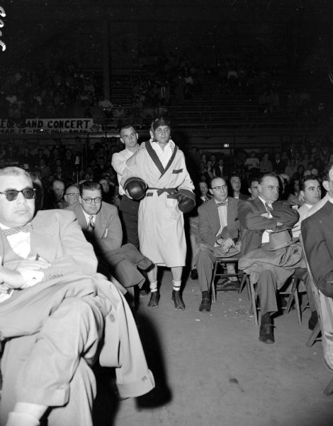 Jim Bernd of New Richmond keeps Bobby Meath's shoulders loose as he heads for the ring. Meath is wearing a light-colored robe, head gear, and boxing gloves. They are walking down an aisle lined with seated, male onlookers dressed in suits. In the background, more fans are sitting in the balcony bleachers.