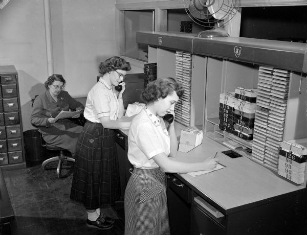 Slightly elevated view of three women in the order department at the American Optical Company, located on 429 State Street. Two women are standing at a counter talking on telephones and taking paper orders. The third woman is sitting in a chair in the background, reviewing paperwork. There is a wooden filing cabinet to the left.