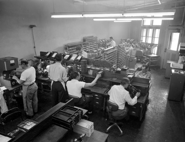 Elevated view of technicians working at the American Optical Company located on 429 State Street. Men are standing and sitting at several work stations to create, grind and adjust glass lenses in a back store room.