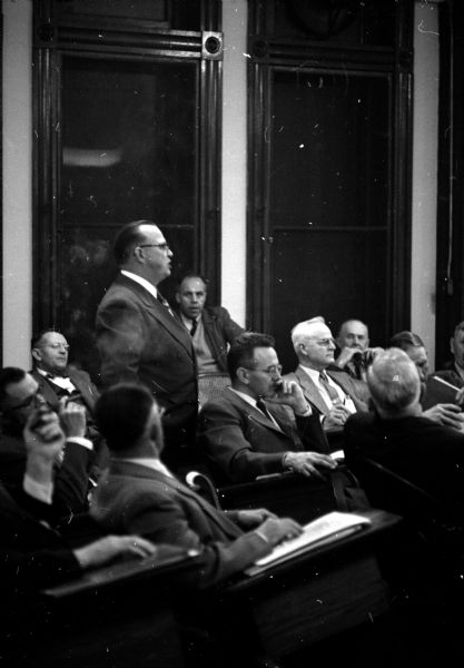 Frank Gugel of Madison Ward 8 standing at his seat to speak as Ivor McBeath of Madison Ward 11 (hand on chin) and Orin McConnell from the Town of Pleasant Springs (right) sit listening.