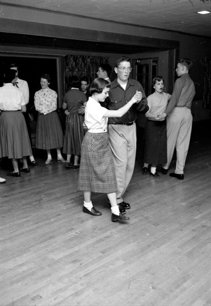 The Shorewood Hills Community League sponsored a dance class for teenagers at the Blackhawk Country Club that was taught by Arthur Murray instructors. Mary Ellen Burns, wearing loafers and white bobby socks, and John Cavanaugh are learning a slow dance, while other teenagers are mingling in the background.