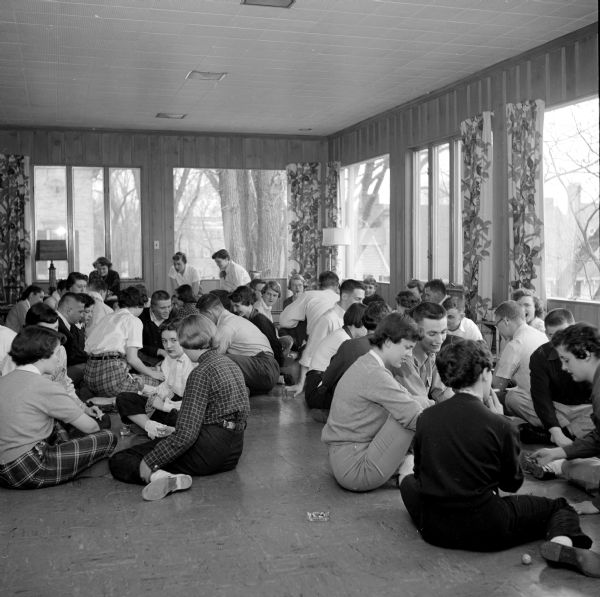 A large group of University of Wisconsin students playing jacks, while sitting on the floor during a tournament at the Alpha Phi House. One  female student is smoking a cigarette.
