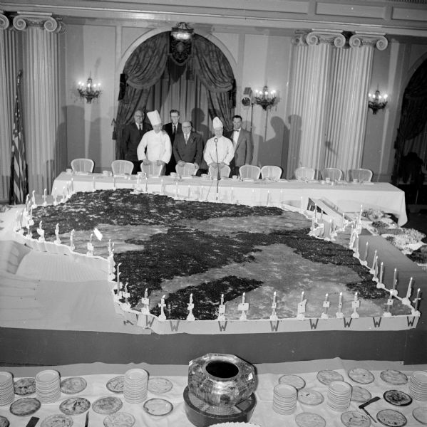 Food from every food-producing county in the state and seventy-one candles - one for each county - frames a 25-foot long map of Wisconsin at the preview event for the 1954 edition of <i>This Is Wisconsin</i> booklet, published by the Wisconsin Tourist Bureau. The event was held at the Hotel Loraine. A group of men with two chefs are standing behind the head table, and a banquet table in the foreground has stacked small plates.