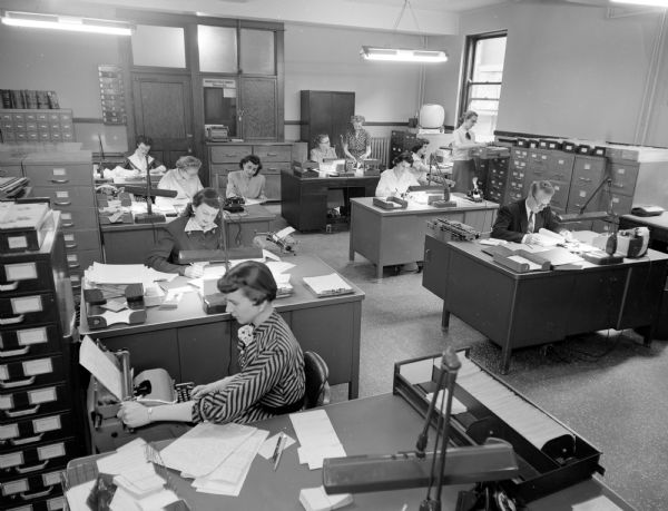 Civilian employees in the Madison police department records center. Staff are sitting at their desks in an open office space with different filing cabinets. One woman is using a typewriter, and another woman is checking records in a drawer.