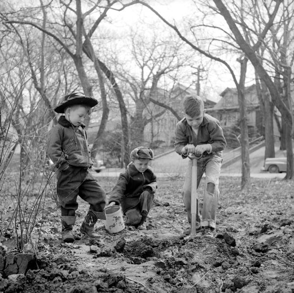 Rickey Holmes, David Dopkins, and Dick Quinlan digging for worms before heading to the Tenney Park lagoon to fish. Dick is using a shovel to overturn wet dirt and David is holding a pail, while crouching. Rickey is looking on while wearing a cowboy hat.