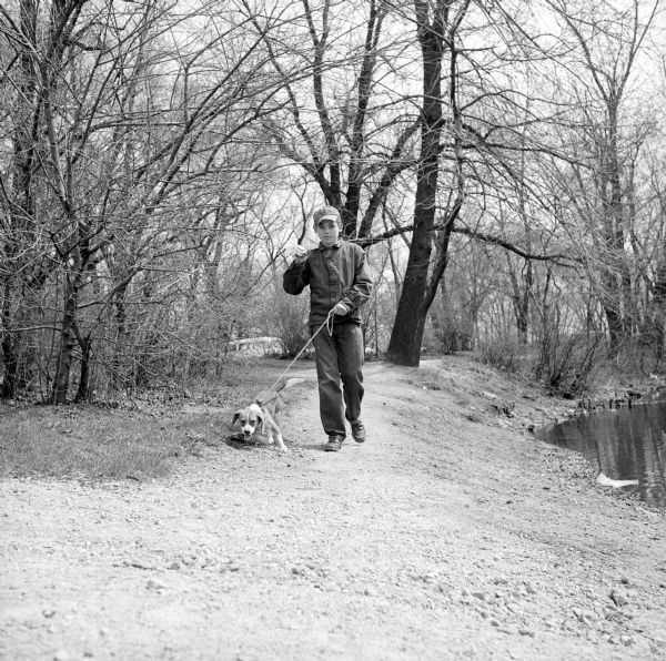Emil Thomas taking his beagle, BeBe, on a leash with him to fish. Walking along the dirt shore of the pond at Tenney Park, the dog is pulling on the leash, while Emil is carrying his fishing rod over his shoulder.