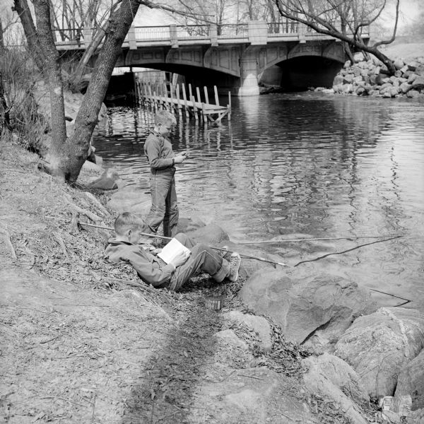 Milwaukee boys, John and David Malec, in Madison to visit their grandmother, Mrs. Louis Malec at 1134 E. Gorham Street. John is fishing on the Yahara River near the Tenney Park locks, while David is reading a book and resting his feet on a rock. The Sherman Avenue bridge and stone riprap are in the background.