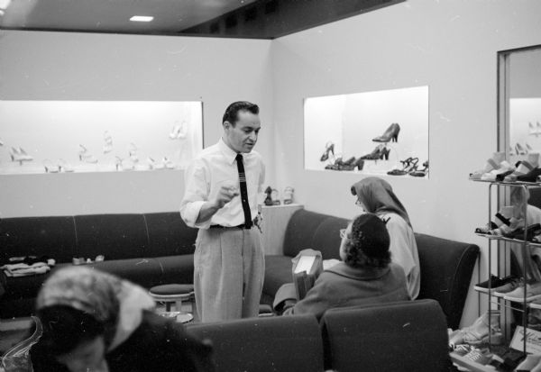 Shoe salesman, Larry Borenstein, answering a woman's questions at the Borenstein Dry Goods Store located at 1980-81 Atwood Avenue. She is sitting on a couch and holding a new, boxed pair of shoes, next to another woman wearing a scarf sitting on a diagonal couch. Shoes are on racks and in recessed wall-displays.