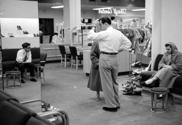 Shoe salesman, Larry Borenstein, assisting a customer at Borenstein's Dry Goods Store located at 1980-81 Atwood Avenue. A young man, perhaps a sales assistant, is seen reflected in a full-length mirror on the far left, and a woman wearing a scarf on her head is sitting and waiting on a couch.