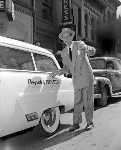 James Roy Miller, photographer for <i>The Capital Times</i>, standing next to a white car advertising "Photography by Edwin Stein, 1417 University Ave." With whitewall tires, it is parked downtown on the  100-block of East Main Street. A car parked behind Stein's car has advertising for "Pete Wilson, Distributor."
 