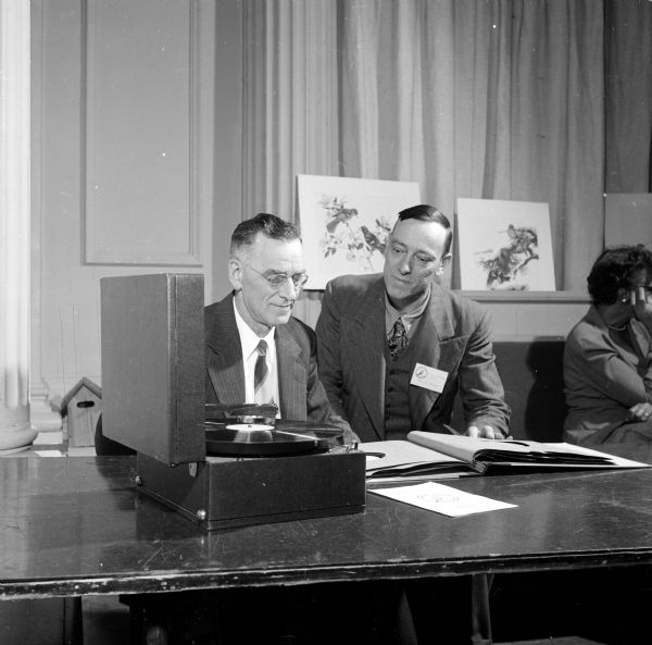 A.L. Throne of Milwaukee and Karl E. Bartel of Blue Island studying bird songs at the reception of the Wisconsin Society for Ornithology at the Memorial Union. They are listening to vinyl records on a portable phonograph. Drawings or paintings of birds are on view behind them.