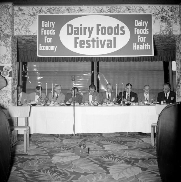 Head table of eight men at the Governor's luncheon for Dairy Week held at the Edgewater Hotel. Walter Kohler, Jr. is seated behind the standing microphone. A banner above them reads: "Dairy Foods For Economy. Dairy Foods Festival. Dairy Foods For Health."